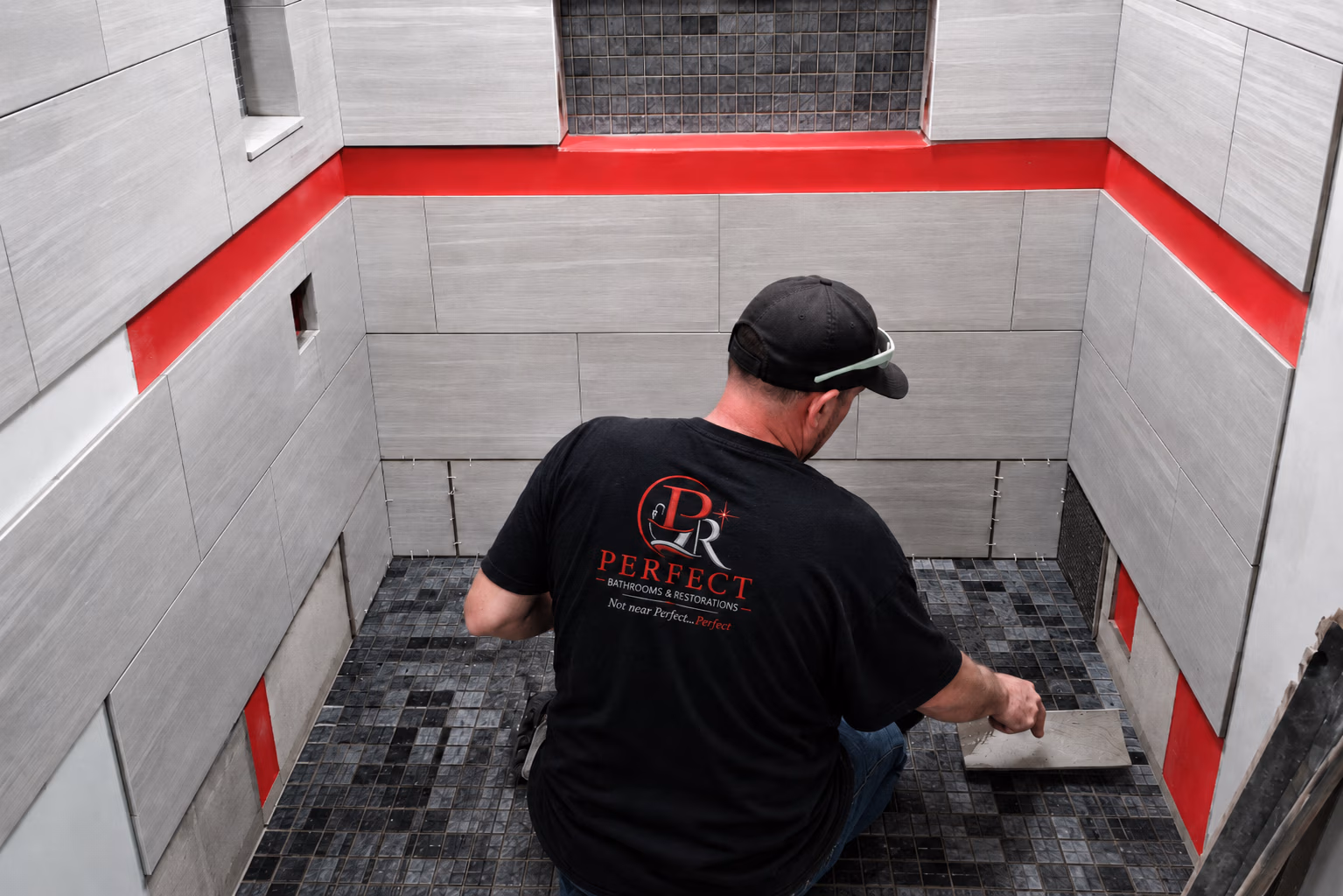 Worker in black shirt tiling a shower floor with dark square mosaic tiles and light wall tiles.