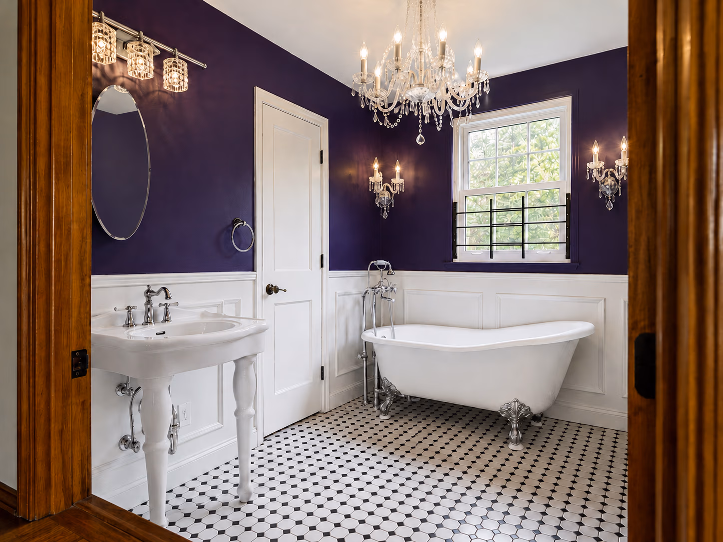 Elegant bathroom with clawfoot tub, pedestal sink, purple walls, white wainscoting, chandelier, and tile floor.