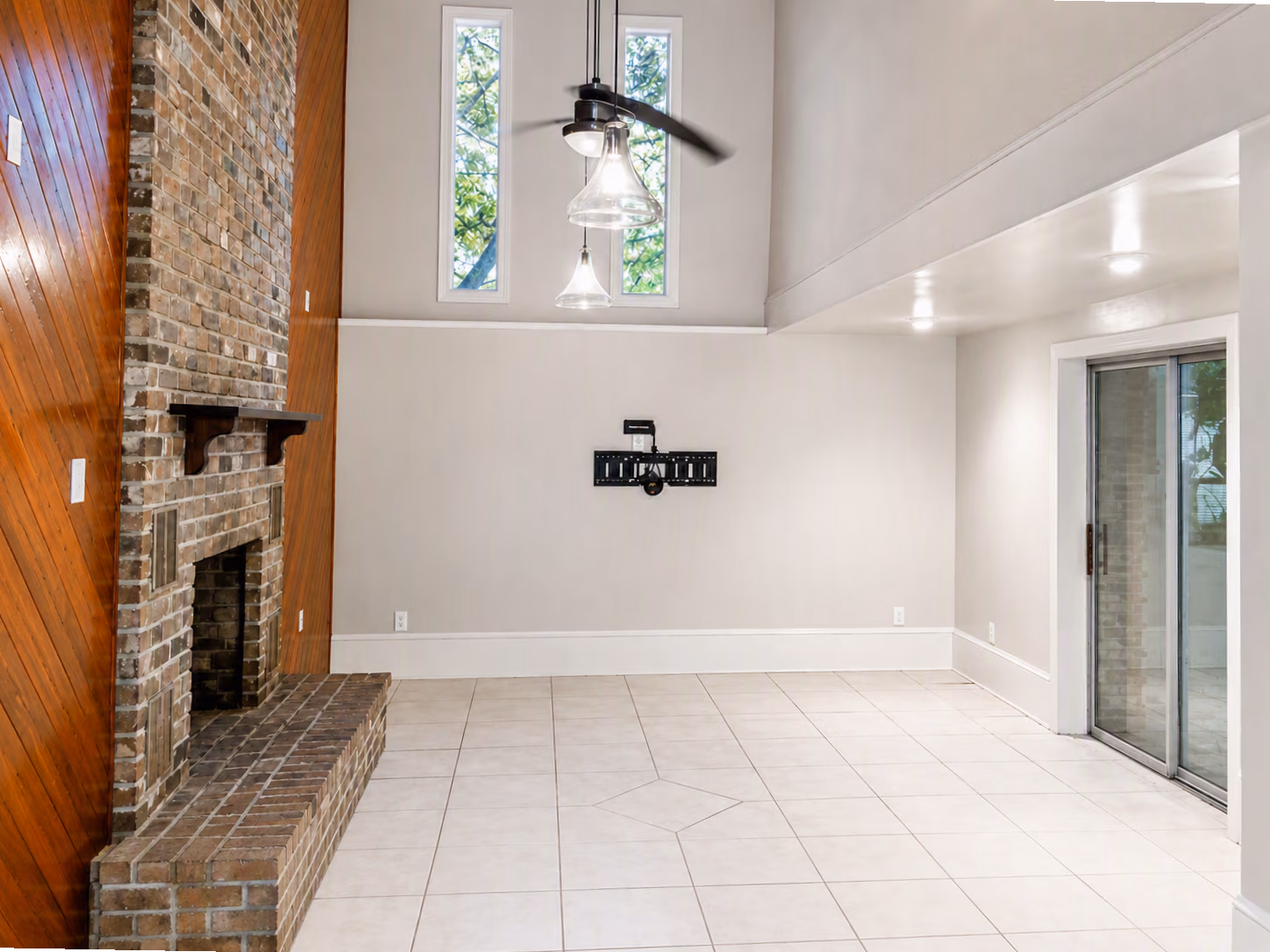 Empty room with brick fireplace, wooden paneling, tiled floor, tall windows, ceiling fan, and sliding glass door.