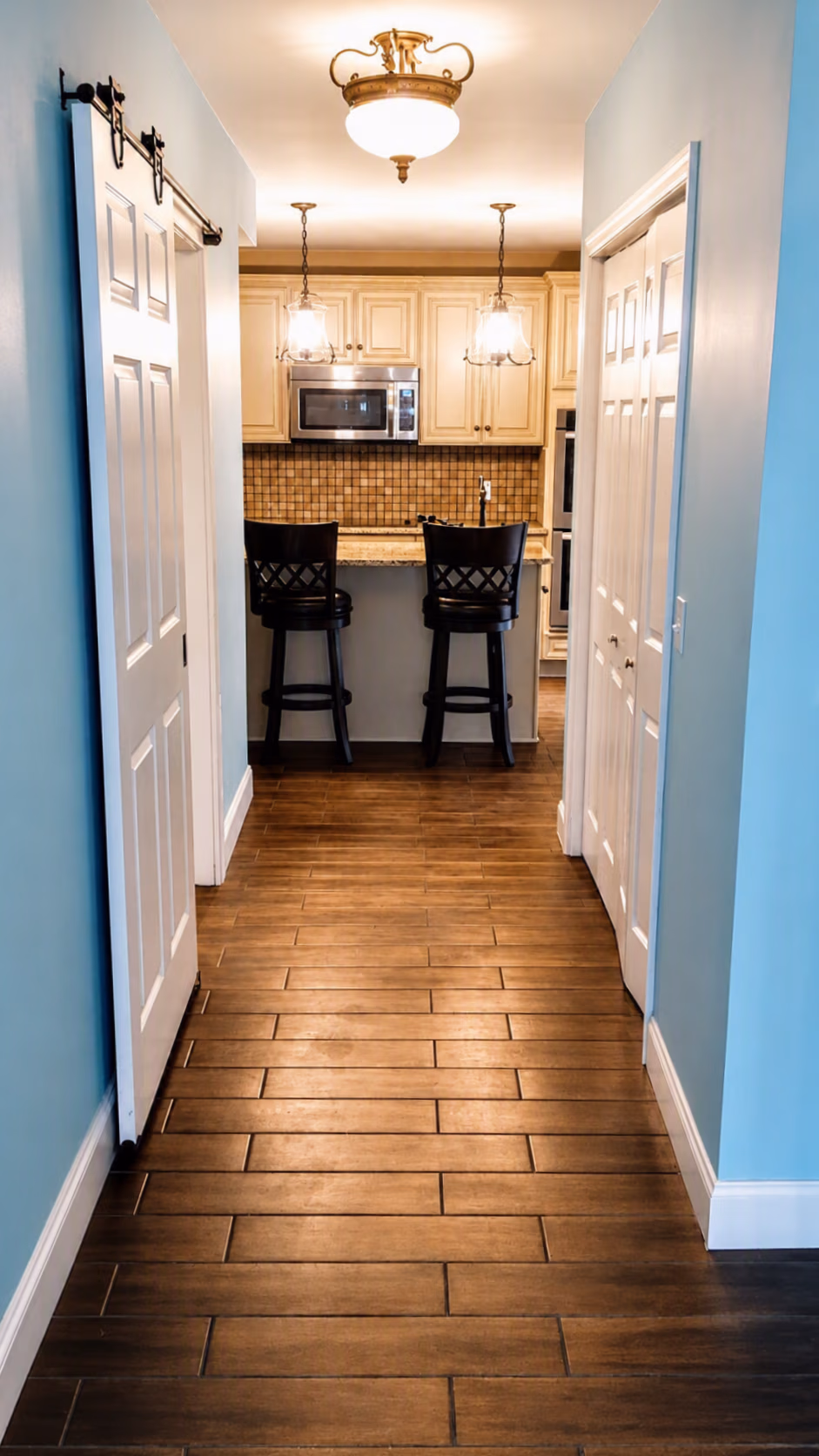 Hallway with wood floor leads to a kitchen with two black stools and beige cabinets.