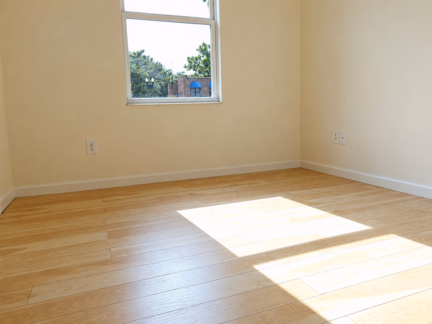 Empty room with light wood floor, beige walls, and a window letting sunlight in.